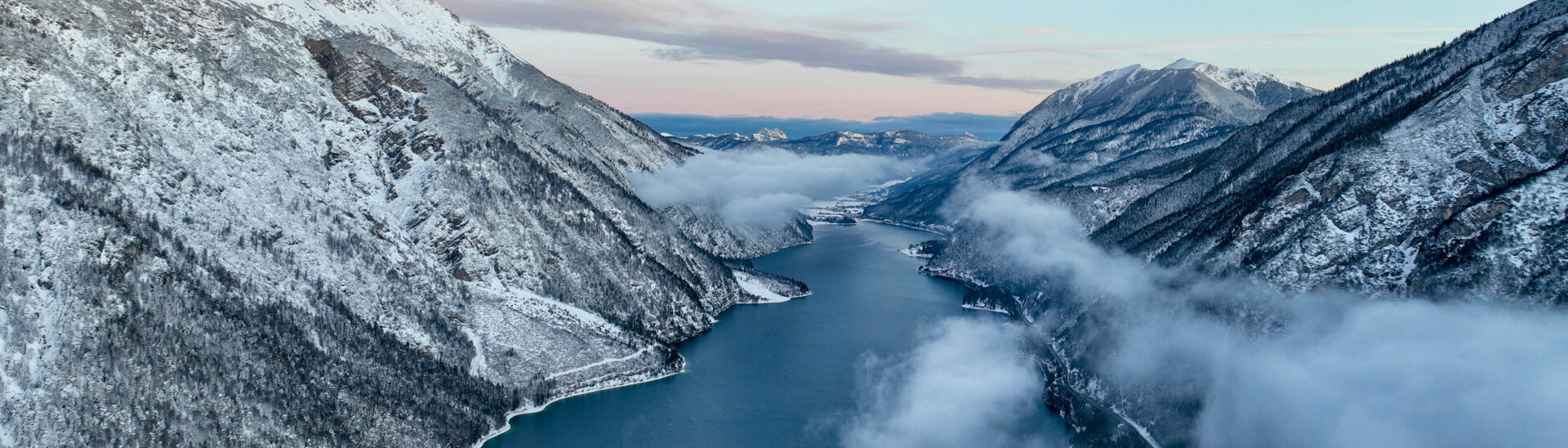 Winter in der Region Achensee Eine atemberaubende Winterlandschaft zeigt schneebedeckte Berge, die an dem ruhigen, blauen Achensee liegen. Nebelschwaden ziehen über das Wasser und zwischen den Bergen, während der Himmel in sanften Pastellfarben erscheint.