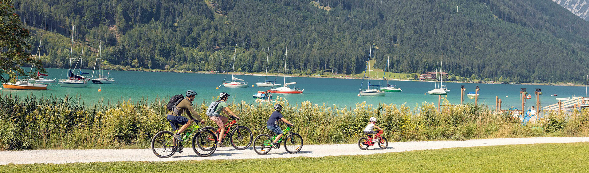 A family with children explores the lakeshore in Maurach am Achensee by bike. In the background of this photo are several sailboats.