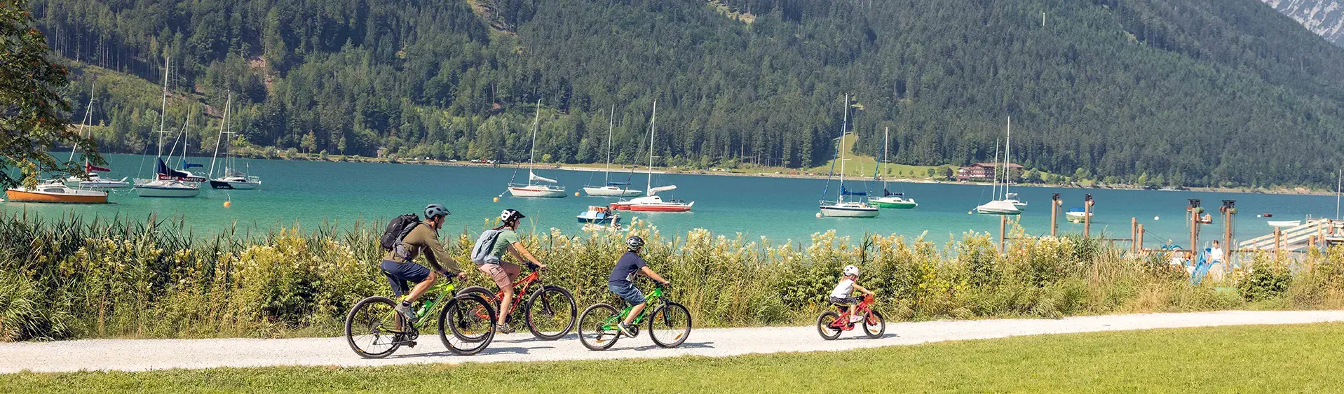 Biken mit der Familie in Maurach am Achensee Einen Familienausflug mit dem Rad am Seeufer in Maurach am Achensee machen. Im Hintergrund sind zahlreiche Segelboote zu sehen.