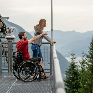 Ein Mann im Rollstuhl und seine Freundin genießen den Ausblick vom Zwölferkopf auf die Region Achensee im Sommer..