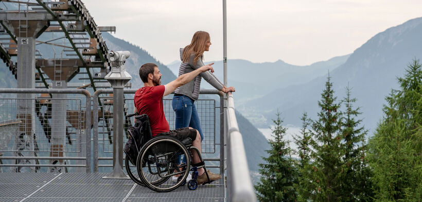 Ein Mann im Rollstuhl und seine Freundin genießen den Ausblick vom Zwölferkopf auf die Region Achensee im Sommer..
