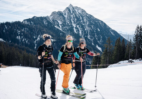 Three female ski tourers stand on a groomed piste in the Rofan mountains. They are dressed in colorful ski gear and are smiling as they navigate the terrain. The atmosphere is lively and energetic, highlighting their enjoyment of the winter sports.