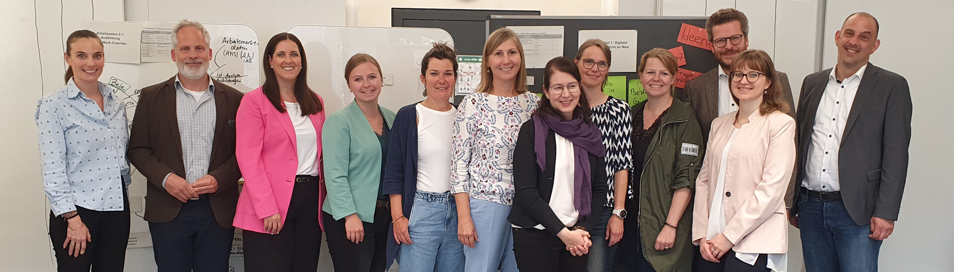 Ein Gruppenfoto von dreizehn New Work Teammitgliedern in einem hellen Raum. Sie lächeln in die Kamera. Im Hintergrund sind Flipcharts und Zeitplanungstafeln sichtbar. 