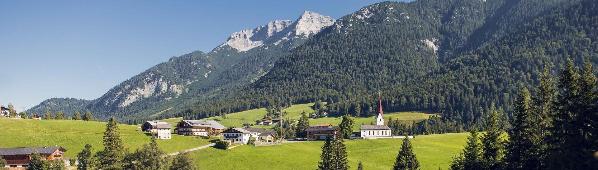 Blick auf das Bergsteigerdorf Steinberg am Rofan, eingebettet in eine sattgrüne, sanft hügelige Landschaft. Rund um den Ort erheben sich imposante Berggipfel und prägen das alpine Panorama.