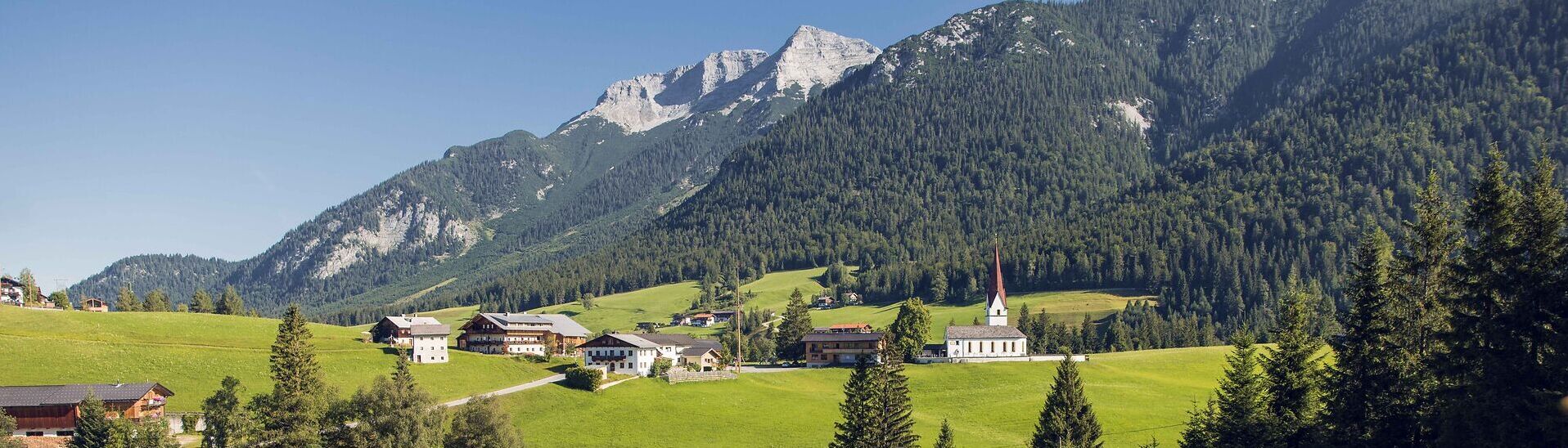 Blick auf das Bergsteigerdorf Steinberg am Rofan, eingebettet in eine sattgrüne, sanft hügelige Landschaft. Rund um den Ort erheben sich imposante Berggipfel und prägen das alpine Panorama.