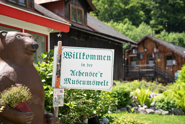 Eine Bärenfigur hält ein Willkommensschild zur Achenseer Museumswelt. Im Hintergrund sind die Museumsgebäude mit Holzbauten zu sehen, umgeben von einem gepflegten Garten und dichtem Wald.