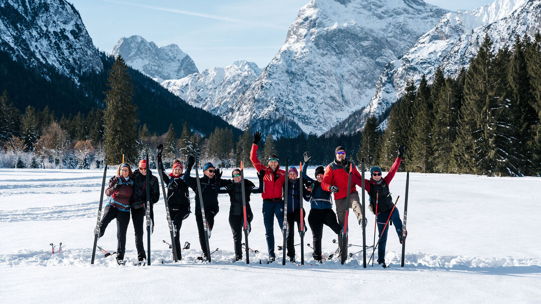 Achensee Langlaufcamp Eine Gruppe von zehn Menschen steht mit Langlaufskiern auf einer verschneiten Loipe vor einer beeindruckenden Bergkulisse am Achensee. Die Teilnehmenden tragen Winter- und Langlaufbekleidung, lächeln in die Kamera und heben fröhlich die Arme oder ein Bein. Im Hintergrund ragen schneebedeckte Gipfel und dunkle Nadelwälder in den klaren blauen Winterhimmel. Das Bild vermittelt Gemeinschaft, Bewegung und die winterliche Atmosphäre des Achensee Langlaufcamps.