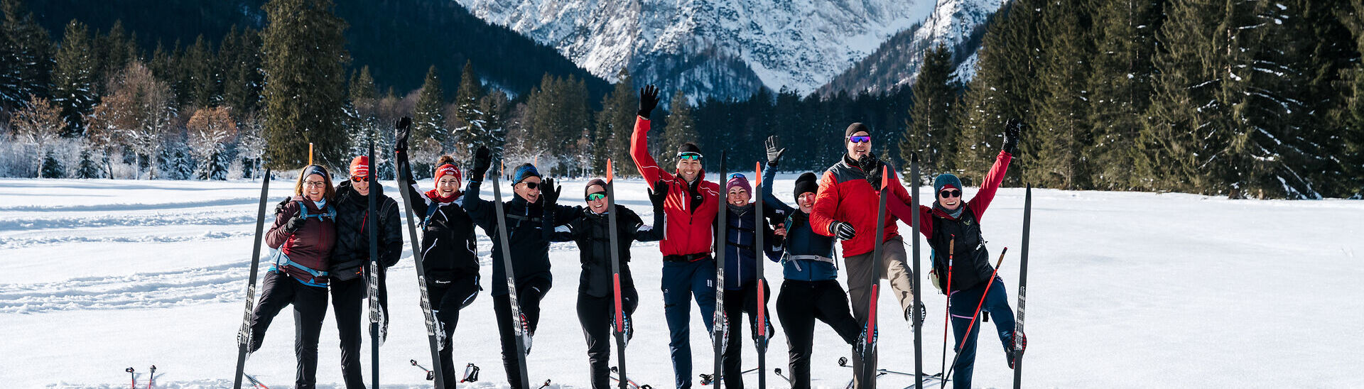 Achensee Langlaufcamp Eine Gruppe von zehn Menschen steht mit Langlaufskiern auf einer verschneiten Loipe vor einer beeindruckenden Bergkulisse am Achensee. Die Teilnehmenden tragen Winter- und Langlaufbekleidung, lächeln in die Kamera und heben fröhlich die Arme oder ein Bein. Im Hintergrund ragen schneebedeckte Gipfel und dunkle Nadelwälder in den klaren blauen Winterhimmel. Das Bild vermittelt Gemeinschaft, Bewegung und die winterliche Atmosphäre des Achensee Langlaufcamps.