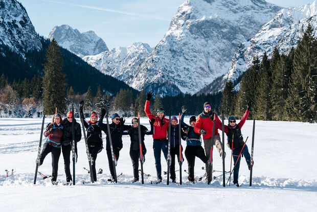 Achensee Langlaufcamp Eine Gruppe von zehn Menschen steht mit Langlaufskiern auf einer verschneiten Loipe vor einer beeindruckenden Bergkulisse am Achensee. Die Teilnehmenden tragen Winter- und Langlaufbekleidung, lächeln in die Kamera und heben fröhlich die Arme oder ein Bein. Im Hintergrund ragen schneebedeckte Gipfel und dunkle Nadelwälder in den klaren blauen Winterhimmel. Das Bild vermittelt Gemeinschaft, Bewegung und die winterliche Atmosphäre des Achensee Langlaufcamps.