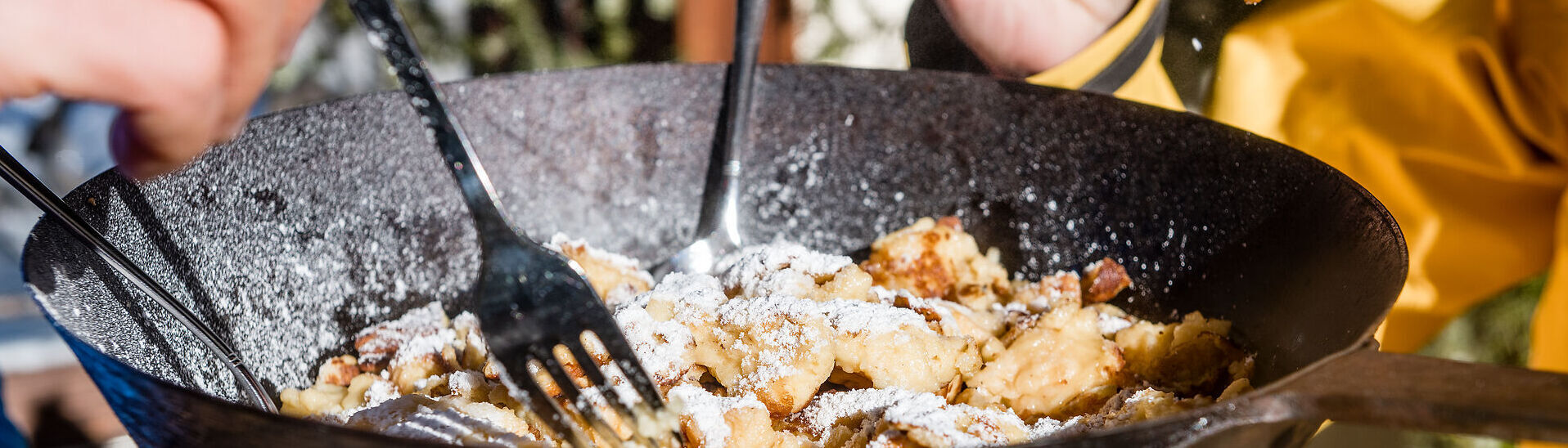 Kulinarischer Genuss am Achensee Mehrere Personen essen gemeinsam Kaiserschmarrn aus einer großen schwarzen Pfanne am Achensee. Mit Gabeln teilen sie die mit Puderzucker bestäubte Süßspeise direkt am Tisch. Die Aufnahme zeigt einen geselligen Moment im Freien bei sonnigem Wetter und vermittelt Genuss, Gemeinschaft und regionale Kulinarik.