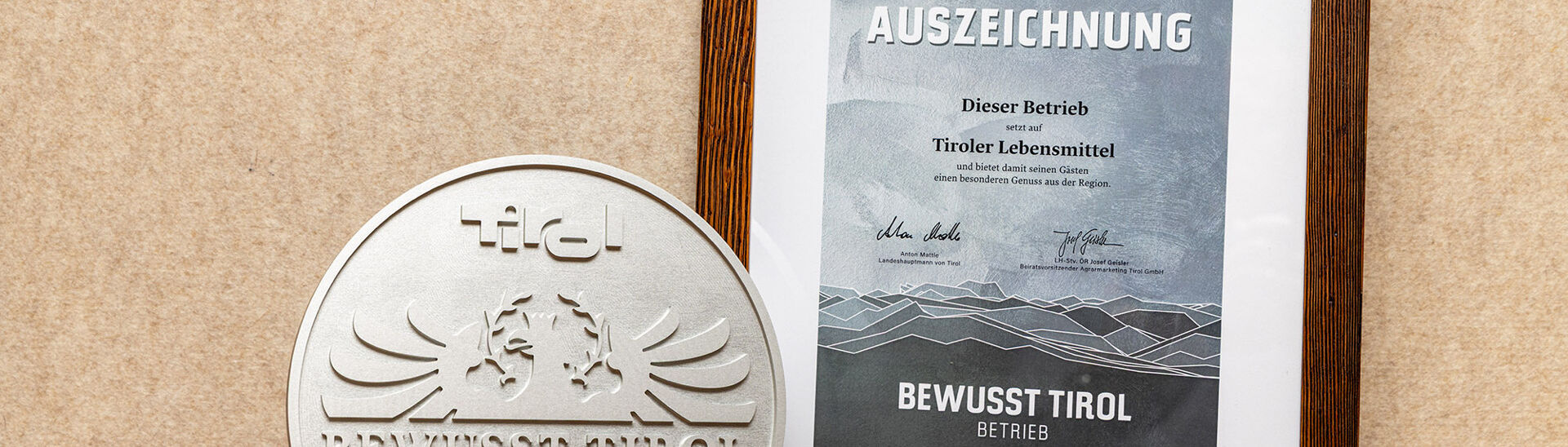 Bewusst Tirol Auszeichnung Ein silbernes Qualitätssiegel mit dem Schriftzug "Bewusst Tirol". Daneben ein gerahmtes Zertifikat mit der Aufschrift "2025 AUSZEICHNUNG" und Informationen über die Unterstützung regionaler Lebensmittel.