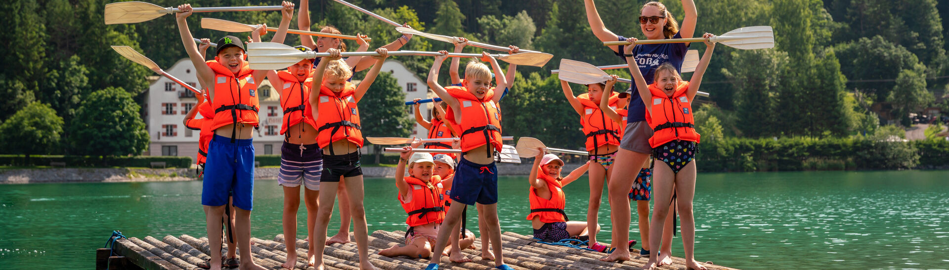 Achensee Children's Programme A group of children wearing bright orange life jackets stand on a wooden raft, joyfully lifting paddles overhead. They are arranged in two rows, with some sitting and others standing, all smiling and celebrating on the shimmering water amidst greenery.