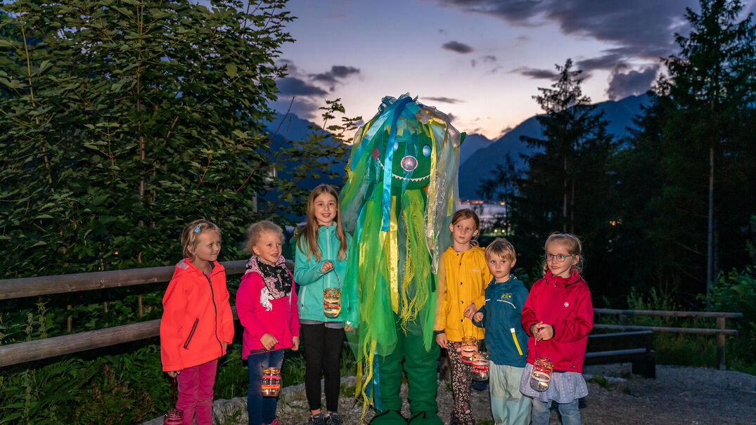 Das Foto zeigt eine Gruppe von sechs Kindern auf einem Weg im Freien während der Dämmerung am Achensee. Jedes Kind hält eine kleine Laterne mit warmem Licht in der Hand. In der Mitte steht eine große, freundliche Figur eines Wassergeists in Grün- und Blautönen mit langen Stoffbändern, einem lächelnden Gesicht und leuchtenden Augen. Die Kinder tragen farbenfrohe Outdoor-Kleidung und stehen eng beieinander. Hinter ihnen erstrecken sich Bäume, Berge und ein Abendhimmel mit dunklen Wolken und den letzten Lichtfarben des Sonnenuntergangs. Die Szene wirkt ruhig, geheimnisvoll und zugleich fröhlich und vermittelt die Atmosphäre einer familienfreundlichen Laternen- oder Sternenwanderung.