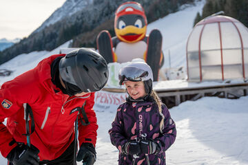 Ein Skilehrer erkundigt sich nach dem Wohlbefinden der kleinen Kursteilnehmerin bei den Planberg- & Wiesenliften in Pertisau. Im Hintergrund Pinguin BOBO.
