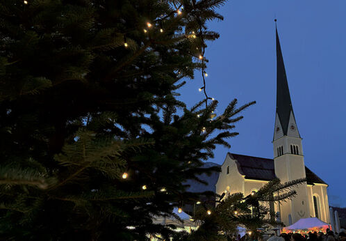 Ein weihnachtlich geschmückter Nadelbaum und im Hintergrund sieht man die Wiesinger Kirche vor dem blauen Abendhimmel.