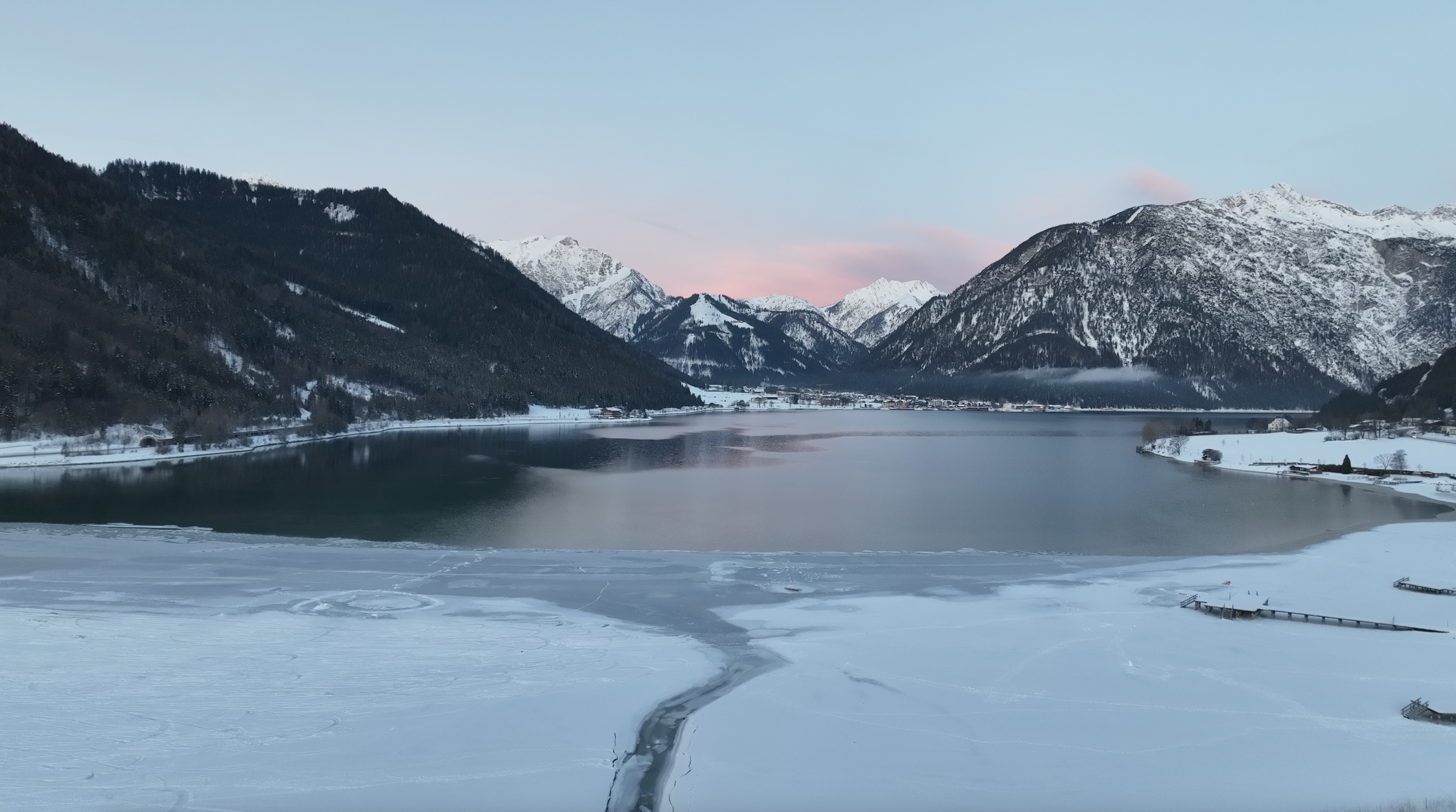 Der Blick zeigt die ruhige Winterlandschaft am Südufer des Achensees mit einem teilweise zugefrorenen See, umgeben von schneebedeckten Bergen. Der Himmel ist blau mit zarten rosa Wolken.